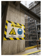 Concrete construction-site pillar with a yellow/black striped warning sign showing a falling-objects hazard and a blue helmet symbol, Czech text warns to wear head protection.