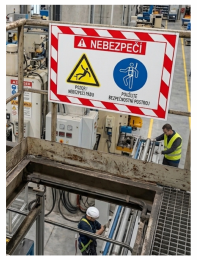 Factory floor with a large danger sign reading 'NEBEZPECÍ' and safety icons; a worker in a hi-vis vest nearby (hazard area).