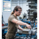 Machinist in safety goggles operates a drill press, clamping a metal block in a vise in a metalworking shop.