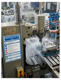 Industrial milling machine in a workshop with a worker using the control wheel; coolant spray and a metal workpiece clamped on the table, safety signage visible on the machine