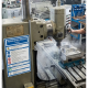 Industrial milling machine in a workshop with a worker using the control wheel; coolant spray and a metal workpiece clamped on the table, safety signage visible on the machine