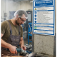 Worker in safety gear (hair net, goggles, apron) grinds a metal piece, sparks flying in a factory setting beside a blue safety sign.