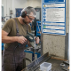 Worker in safety gear drills metal piece on a workbench in a machine shop.