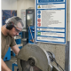 Worker wearing earmuffs and a mesh cap operating a metal-cutting circular saw in a workshop; safety poster on the wall behind him.