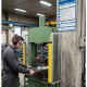 Factory worker wearing ear protection operating a green hydraulic press with a metal workpiece clamped on the die.