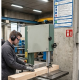 Worker in safety gear uses a vertical bandsaw to cut a wooden beam in a factory workshop.